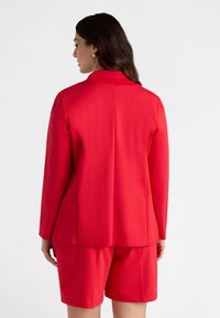 Blazer rojo combinado con pantalones cortos a juego. El blazer cuenta con un cuello y hombros estructurados. Textura suave, corte ajustado y líneas limpias.