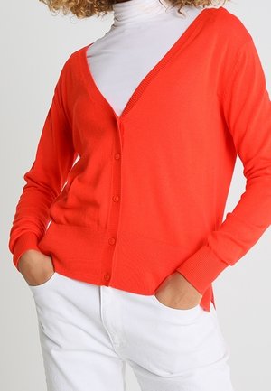 Roter Strickjacke mit tiefem V-Ausschnitt und Knopfverschluss, mit gerippten Bündchen und Saum; getragen über einem weißen Rollkragenpullover und kombiniert mit weißen Hosen.