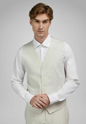 Junger Mann mit hellbraunen Haaren, der ein weißes Hemd und eine cremefarbene, geknöpfte Weste vor einem schlichten grauen Hintergrund trägt.