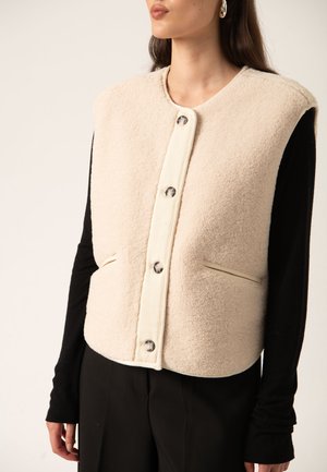 Woman wears cream-colored, buttoned, sleeveless fleece vest over long-sleeve black top and black pants, shown from shoulders to hips.