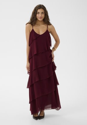 Femeie într-o rochie maxi bordo fără mâneci, cu volane, cu bretele subțiri, stând în picioare și zâmbind, purtând pantofi negri cu vârf ascuțit și o brățară aurie.