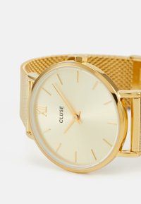 Montre-bracelet en maille dorée avec un cadran rond, un cadran doré clair, le chiffre romain III, des repères horaires simples et un profil fin. Marque : Cluse.