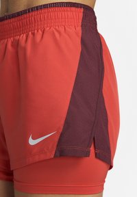 Pantalones cortos de running Nike en rojo y marrón con pantalones cortos de compresión integrados y una cinturilla elástica, ajustados a la cintura y parte superior del muslo de una persona.