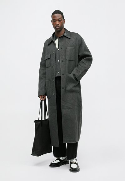 Pelēks vilnas mētelis ar lielām kabatām, melns ādas apkakle, valkāts virs gaiša krekliņa ar pogām, kopā ar melniem biksēm un spīdīgiem mokasīniem.