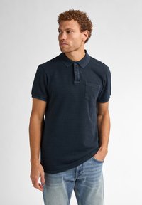 Cămașă polo bleu marin cu design texturat, placket cu două butoane și buzunar pe piept. Mâneci scurte și guler, asociată cu blugi deschiși.