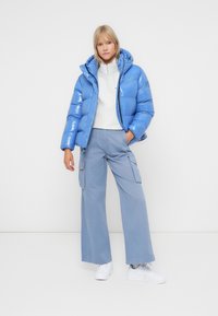 Glanzende blauwe pufferjack met een capuchon, gevuld met isolatie. Gecombineerd met lichtblauwe cargobroek en witte sneakers. Soepele textuur.