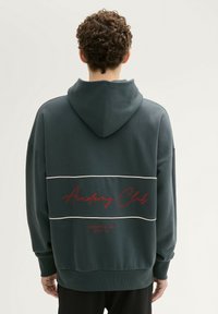 Ciemnozielona bluza z kapturem z przednią kieszenią typu kangur, białymi poziomymi paskami oraz napisem "Academy Club" na plecach w kolorze czerwonym. Miękka tekstura materiału.