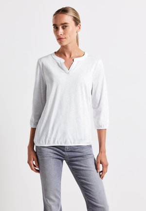 Weißes Langarmshirt mit V-Ausschnitt, leichtem Stoff und gerafften Bündchen, kombiniert mit hellgrauen Jeans. Einfaches und minimalistisches Design.