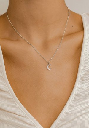 Silberne Halskette mit einer zarten Kette und einem Anhänger in Form des Buchstabens "C", verziert mit kleinen Edelsteinen, die auf nackter Haut und einem leichten Stoffoberteil liegt.