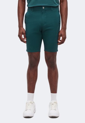 Homme portant un short bleu sarcelle, des chaussettes blanches et des baskets blanches, debout avec les bras détendus le long du corps.