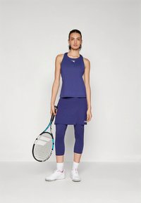 Lila sportos szett, mely ujjatlan felsőből, hozzáillő szoknyából és leggingsből áll, sima anyagból készült, teniszütővel párosítva.