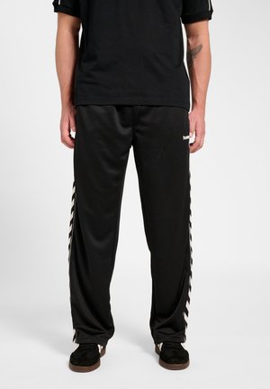 Mand iført sorte træningsbukser med hvide chevron-striber ned langs siderne, kombineret med sorte sko og en sort kortærmet skjorte.