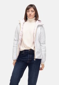 Giacca trapuntata bianca con fodera rosa, cerniera dorata e collo alto, abbinata a un dolcevita color crema e jeans blu scuro.