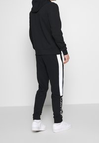 Osoba oblečená v černé mikině s kapucí a černých teplácích s bílými bočními pruhy a bílým textem "Calvin Klein", doplněná o bílé tenisky.