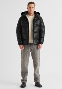 Schwarze, gepolsterte Jacke mit Kapuze, die ein elegantes, glänzendes Finish hat. Getragen über einem cremefarbenen Strickpullover, kombiniert mit grauen Jeans und schwarzen Stiefeln.