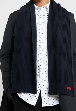 Homme portant une écharpe noire côtelée sur une chemise blanche à pois noirs et un manteau noir, mains dans les poches, étiquette logo rouge sur l'ourlet de l'écharpe.