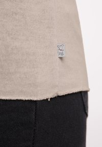 Beige gebreide top met een textuurafwerking, voorzien van een rafelige zoom en een klein logo-label met een afbeelding van een hond. Draagt met zwarte broek.
