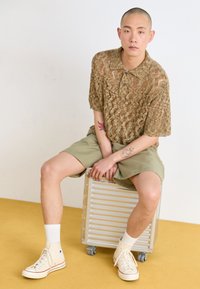 Knit short-sleeve polo shirt in beige, khaki shorts, and beige high-top sneakers with red accents. Sitting on a silver, wheeled storage case.