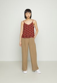 Camisola vermelha com padrão e alças finas, apresentando um design semelhante a folhas. Combinada com calças largas bege e ténis brancos.