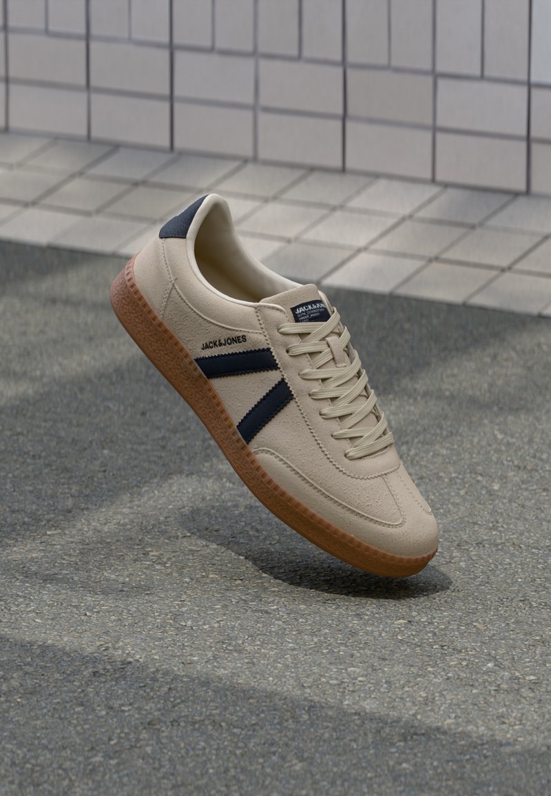 Beige Sneaker mit strukturierter Oberfläche, dunkelblauen Streifen und Gummisohle. Verfügt über eine gepolsterte Zunge und ein traditionelles Schnürsystem für einen sicheren Sitz.