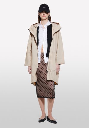 Veste à capuche beige avec une doublure intérieure noire, chemise blanche à boutons, jupe noire en voile à pois et ballerines noires. Une casquette noire complète le look.