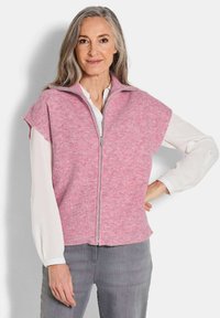 Rosa ärmellose Weste mit hohem Kragen und einem Frontreißverschluss, ausgestattet mit einer weichen, gerippten Textur. Trägt sie über einer weißen Bluse, kombiniert mit grauen Hosen.