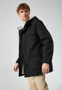 Chaqueta aislante negra con capucha, con forro de forro polar y cuatro bolsillos frontales. Diseñada con una cremallera frontal y cuerdas ajustables.
