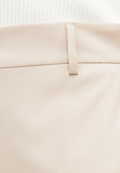 Κοντινή λήψη στη μέση μπεζ παντελονιού με θηλιά ζώνης, φορεμένη πάνω από λευκό ριμπ ύφασμα.
