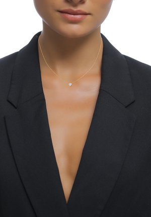 Gros plan d'une personne portant un collier fin en or avec un pendentif en diamant en forme de cœur et un blazer noir avec un décolleté plongeant.