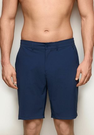 Mann trägt marineblaue knielange Shorts, steht vor einem schlichten hellen Hintergrund, sichtbar von der Taille bis zu den Knien.