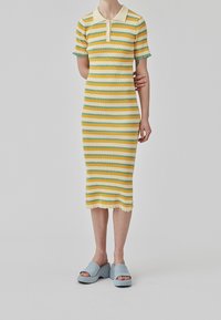 Robe côtelée à rayures jaunes et vertes avec un col polo, des manches courtes et un ourlet effiloché. Portée avec des sandales à plateforme bleues.