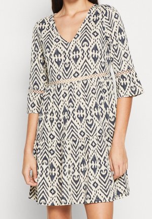 Mujer con vestido beige y negro estampado con escote en V, mangas tres cuartos y detalles de encaje en la cintura y las mangas.