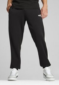 Schwarze Jogginghosen mit weißen Seitenstreifen, ausgestattet mit einem elastischen Bund und Taschen. Kombiniert mit grau-weißen Turnschuhen.