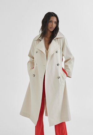 Trench coat beige de doble botonadura con gran cuello, bolsillos laterales forrados en rojo y botones plateados. Tejido suave con un diseño estructurado.