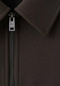 Tkanina o ciemnobrązowym kolorze i fakturze, z gładkim wykończeniem, wyposażona w metalowy zamek błyskawiczny z logo oraz kołnierz w kształcie ostro zakończonym.