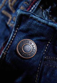 Dżinsowa tkanina zbliżenie na mosiężny guzik z wytłoczonym tekstem "JACK'S SPORTSWEAR" oraz kontrastującymi żółtymi szwami.