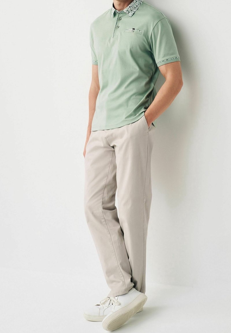 Homme portant un polo vert clair avec un col et des manches fleuris, un pantalon beige et des baskets blanches, debout devant un mur blanc.