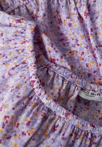 Tissu lilas clair avec de petits motifs éparpillés en rose, orange et blanc. Présente des fronces élastiquées le long des bords et une étiquette d'entretien.