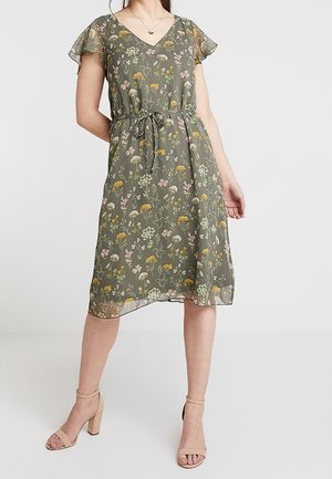 Kobieta ubrana w oliwkową, kwiatową sukienkę do kolan z krótkimi falbaniastymi rękawami i paskiem do wiązania, w połączeniu z beżowymi sandałami na słupku z paskiem wokół kostki.