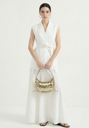 Donna con capelli scuri, indossa un gilet bianco senza maniche con cintura e una gonna lunga, tiene una borsa lucida dorata con paillettes su uno sfondo semplice.