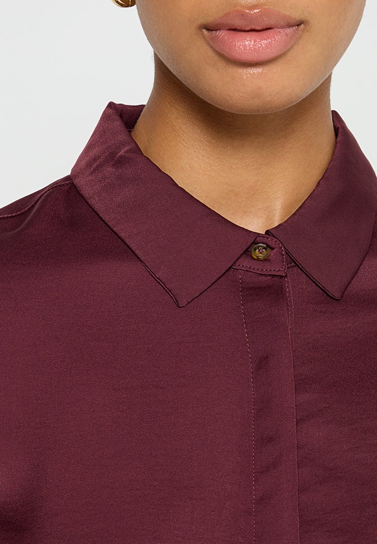 Nahaufnahme von Lippen und Untergesicht, die glänzenden Lippenbalsam tragen und ein zugeknöpftes dunkelviolettes Hemd mit Kragen.