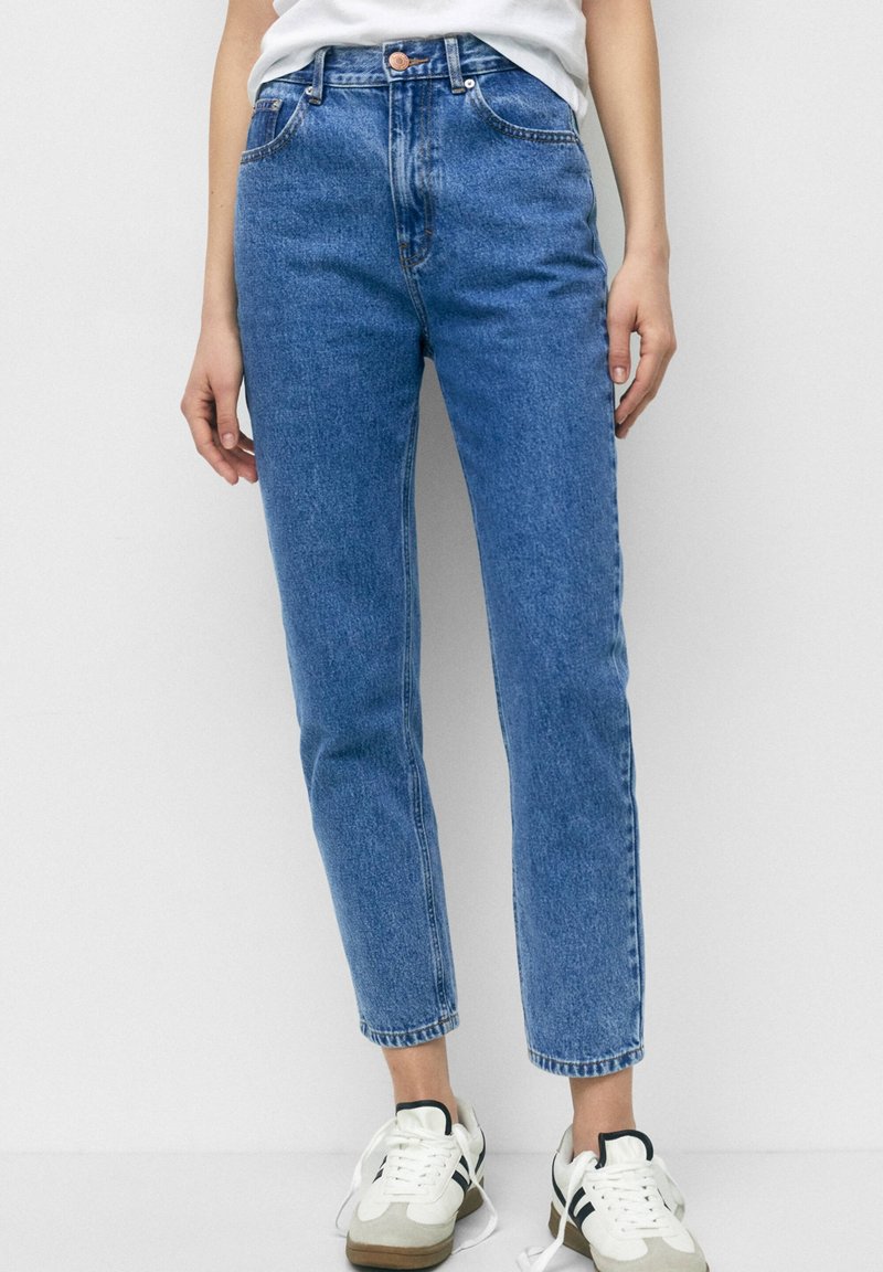 Femme portant un jean bleu taille haute, un t-shirt blanc rentré et des baskets blanches avec des accents noirs, debout devant un fond blanc uni.