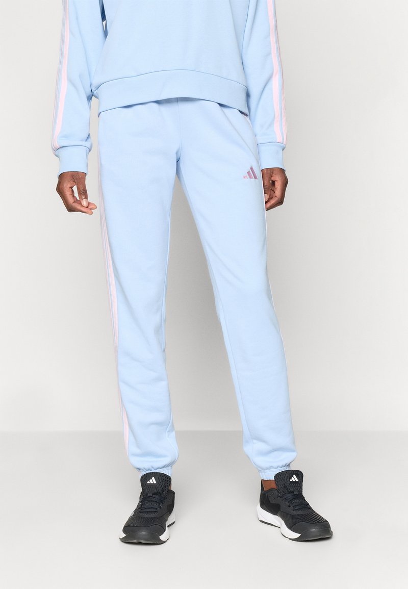 Svetlomodrý teplákový komplet s mikinou na okrúhlym výstrihom a zodpovedajúcimi teplákmi. Na nohaviciach sú tri ružové pruhy. Čierne športové topánky.