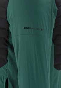 Sezione di manica di una giacca verde e nera con il logo "ENDURANCE" stampato sul tessuto verde.
