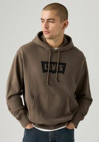 Sudadera de algodón marrón con un logo negro de Levi's, capucha con cordón, bolsillo frontal y puños de canalé. Lleva puesta sobre una camiseta blanca y jeans azules.