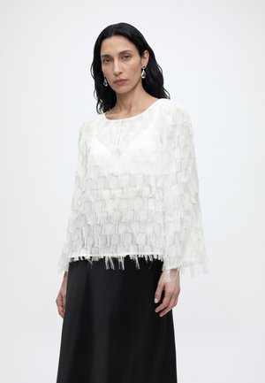 Naine pikkade mustade juustega, kes kannab valget tekstuuriga fringitud pluusi ja musta seelikut, seisab tavalise heleda tausta ees.