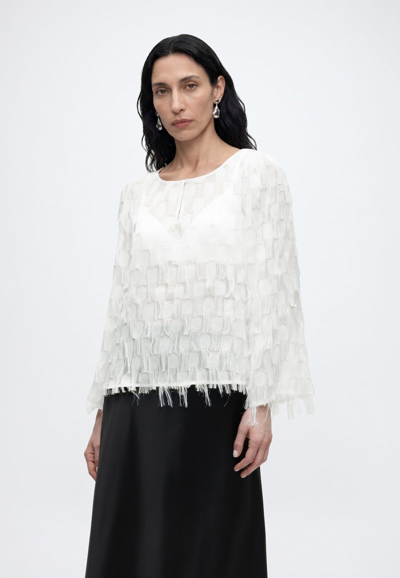 Γυναίκα με μακριά μαύρα μαλλιά που φοράει λευκό υφασμάτινο μπλουζάκι με κρόσσια και μαύρη φούστα, στέκεται μπροστά σε απλό φωτεινό φόντο.