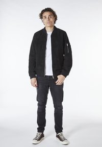 Svart mockabomberjacka med ribbade manschetter och en dragkedjeficka på ärmen, i kombination med slitna mörka jeans och bruna högstövlar.