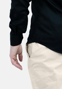 Zwarte longsleeve shirt met knopen op de manchetten, gecombineerd met lichtbeige broek, met een hand nabij de zijzak op een neutrale achtergrond.