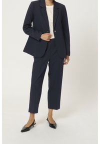 Kobieta w granatowej marynarce i dopasowanych, krótkich spodniach, białej bluzce i czarnych czółenkach na obcasie, stojąca na tle jednolitego tła.